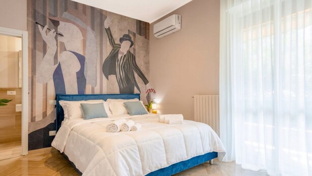 Une chambre à coucher avec un lit, un climatiseur fixé au mur, une fenêtre avec des rideaux légers et un tableau accroché au mur.