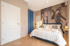 A bedroom with a bed, a door, a wall with a mural, and a nightstand with a lamp.