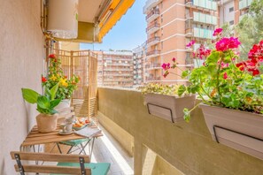 A balcony with potted plants, a table with cups and a plate, and a view of multi-story buildings.