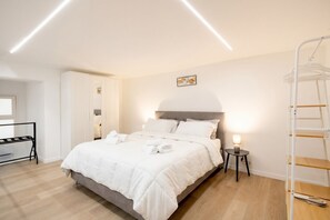 Una cama bien hecha con ropa de cama blanca, una mesita de noche con lámpara, un armario blanco y una escalera adosada a la pared.
