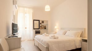 Una habitació d'hotel amb un llit, escriptori amb ordinador, una cadira, taula de nit amb làmpada i finestra amb cortines.