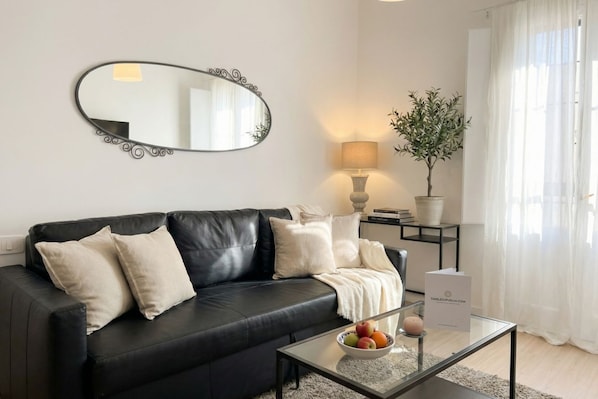 A modern living room with a black leather sofa, a glass coffee table with fruit, a decorative mirror, a lamp, and a potted plant.