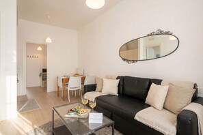 A living room with a black leather sofa, a glass coffee table with snacks and wine glasses, a dining area with a white table and chairs, and a large mirror on the wall.