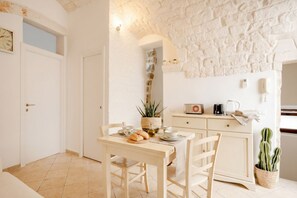 A cozy dining area with a stone wall, a wooden table, chairs, a small plant, a radio, and a kitchen area with a refrigerator. - Mia & Mimi – Accogliente Rifugio nel Centro Storico (Ostuni)