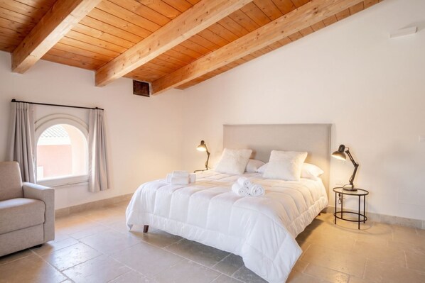 A bedroom with a large bed, two bedside lamps, a chair, and a window with curtains.