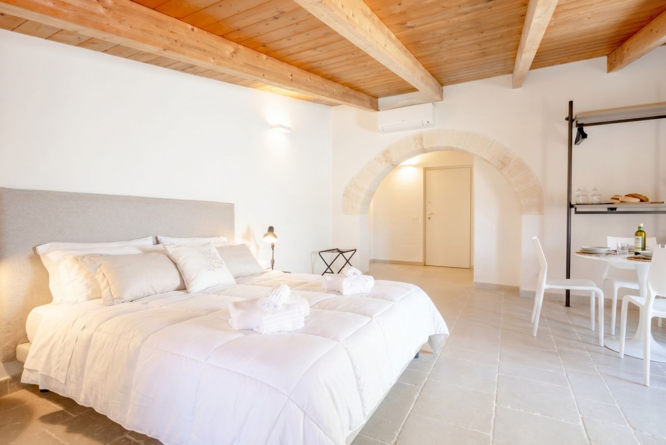 Une chambre dotée d’un grand lit, d’un plafond en bois, d’une arche en pierre et d’un coin repas avec des chaises et une table.