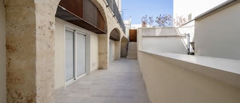 A narrow, tiled corridor with arched doorways leading to a building with a white facade and a balcony.
