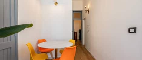 Un couloir étroit avec une porte blanche, une table ronde entourée de chaises orange et une petite plante.