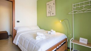 A neatly made bed with white bedding and rolled towels, a bedside lamp, a wall-mounted coat rack, and a small shelf with a sign.