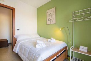 A neatly made bed with white bedding and rolled towels, a bedside lamp, a wall-mounted coat rack, and a small shelf with a sign.