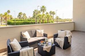 A balcony with wicker furniture, a coffee table with a fruit bowl, and a view of palm trees and greenery. - Sky  Gate  Green  –  Airport  Terrace  Suite (Bari)