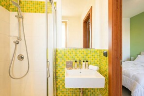 Une salle de bain avec une douche, un lavabo et un lit visible à travers une porte ouverte.