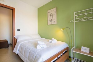 Ein ordentlich gemachtes Bett mit weißer Bettwäsche und aufgerollten Handtüchern, eine Nachttischlampe, ein an der Wand befestigter Kleiderhaken und eine kleine Ablage mit einem Schild.