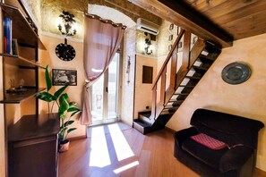 Un salon chaleureux avec un escalier, des poutres en bois, un canapé noir, une plante en pot et une horloge murale.