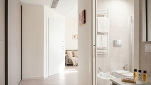 A modern bathroom with a bathtub, a sink, and a mirror. There is a door leading to a bedroom with a bed and a nightstand.