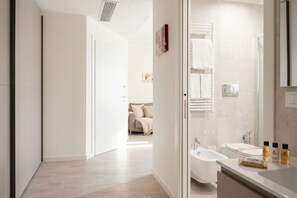 A modern bathroom with a bathtub, a sink, and a mirror. There is a door leading to a bedroom with a bed and a nightstand.