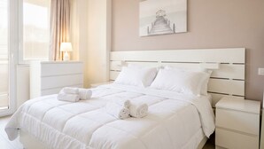 A hotel room with a large bed, a bedside table, a lamp, a window with curtains, and a painting on the wall.