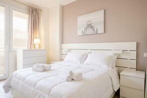 A hotel room with a large bed, a bedside table, a lamp, a window with curtains, and a painting on the wall.