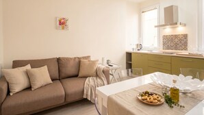A modern living room with a sofa, a dining table with a plate of olives and a bottle of oil, and a kitchen area with a window and a decorative backsplash.