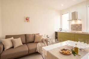 A modern living room with a sofa, a dining table with a plate of olives and a bottle of oil, and a kitchen area with a window and a decorative backsplash.