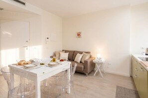 A modern, well-lit room with a dining table set for two, a sofa, and a small kitchen area. - Suite Calle - City Center Modern Apartment+Balcony (Bari)