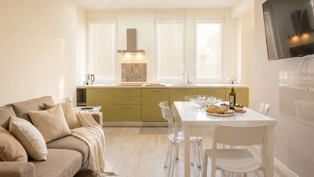 A modern living room with a sofa, a dining table with chairs, a kitchen area with a stove, and a TV mounted on the wall.