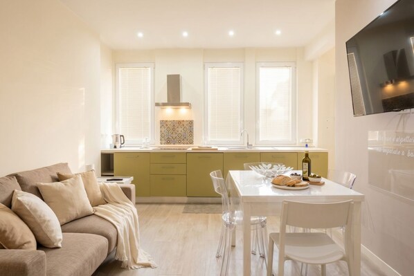 A modern living room with a sofa, a dining table with chairs, a kitchen area with a stove, and a TV mounted on the wall.