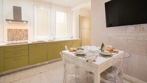 A modern kitchen with a dining area, featuring a white table, clear chairs, and a flat-screen TV mounted on the wall.