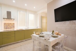 A modern kitchen with a dining area, featuring a white table, clear chairs, and a flat-screen TV mounted on the wall.