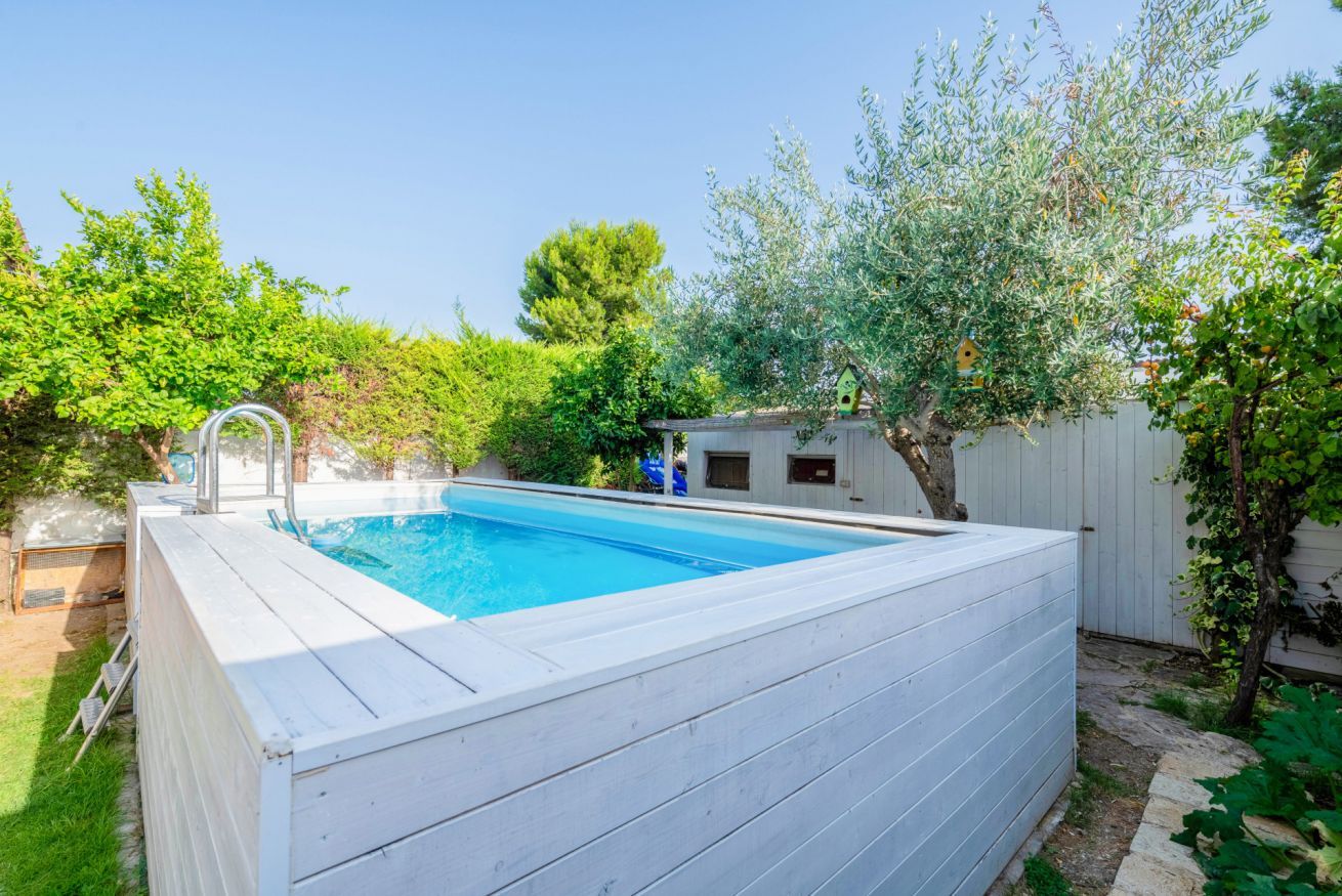 Una piscina rettangolare con bordo in legno, circondata da vegetazione lussureggiante e una recinzione bianca.