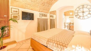 Quarto com teto de pedra, cama com roupa de cama estampada, relógio e área de jantar visível por um arco.