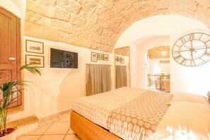 A bedroom with a stone ceiling, a bed with patterned bedding, a clock, and a dining area visible through an archway.