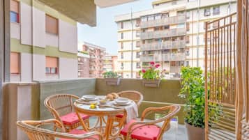 Un balcón con sillas de mimbre y una mesa lista para comer, rodeado de plantas en macetas y vistas a un edificio residencial.