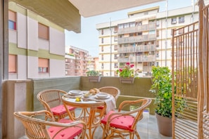 A balcony with wicker chairs and a table set for dining, surrounded by potted plants and overlooking a residential building.