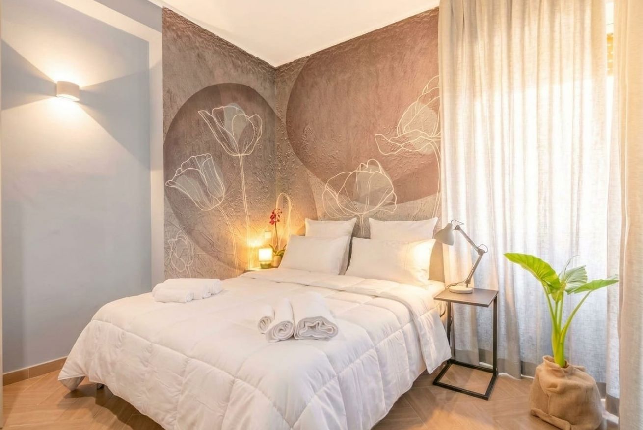 Une chambre à coucher avec un lit, des oreillers, une table de chevet avec une lampe, un mur orné d’un motif floral et une plante.