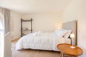 Une chambre d’hôtel bien rangée, avec un lit, une table de chevet dotée d’une lampe, un plancher en bois et une fenêtre avec des rideaux.