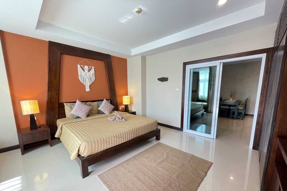 A modern bedroom with a large bed, two bedside lamps, a decorative wall hanging, and a view into a dining area through a glass door.
