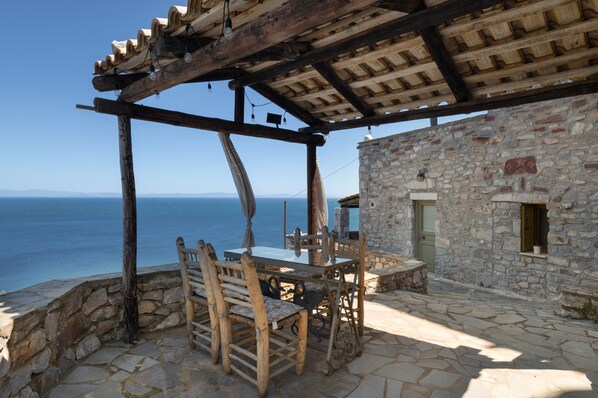 A stone building with a wooden pergola, a table with chairs, and a view of the sea.