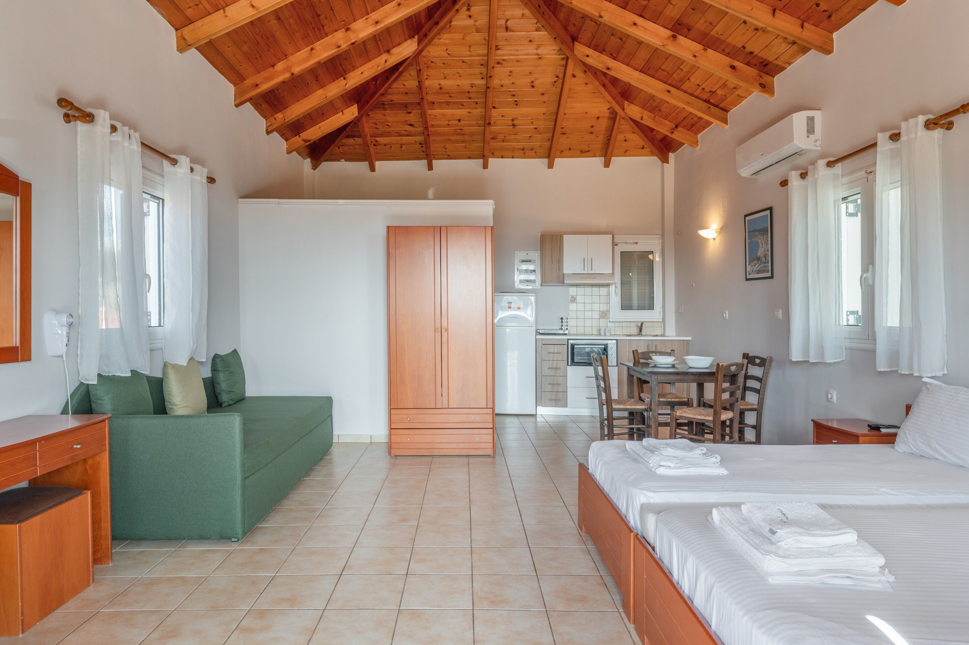 Quarto com teto de madeira, sofá verde, cama com toalhas, cozinha e mesa de jantar.