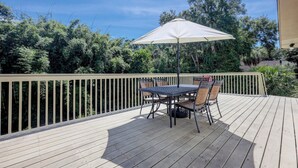 Una terraza de madera con una mesa y sillas bajo un paraguas blanco, rodeada por una barandilla de madera y abundante vegetación.