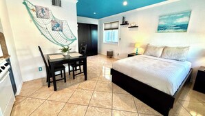 A bedroom with a bed, a desk with a chair, a dining table, and a kitchen area.