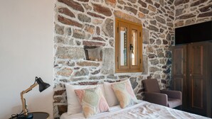 A bedroom with a stone wall, a bed with patterned pillows, a chair, a desk with a lamp, and a wardrobe. - Apartment 'Castle Rizaraki 206' with Sea View, Wi-Fi and Air Conditioning (Γύθειο)