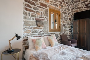A bedroom with a stone wall, a bed with patterned pillows, a chair, a desk with a lamp, and a wardrobe.