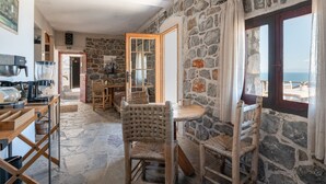 A stone-walled room with a dining table and chairs, a kitchen area with a coffee machine, and a view through a window. - Apartment 'Castle Rizaraki 206' with Sea View, Wi-Fi and Air Conditioning (Γύθειο)