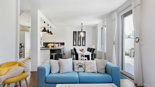 A modern living room with a blue sofa, yellow chair, and a dining area with a kitchen in the background.