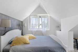 Ein Schlafzimmer mit schrägen Wänden, einem Bett mit grau-gelber Bettdecke, zwei Nachttischlampen und einem Fenster mit durchsichtigen Vorhängen.