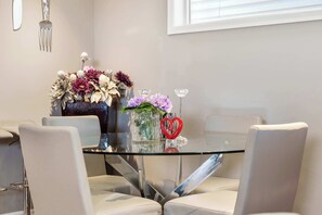Een eethoek met een glazen tafel, omringd door vier stoelen, een vaas met bloemen en een decoratieve hartvormige decoratie.