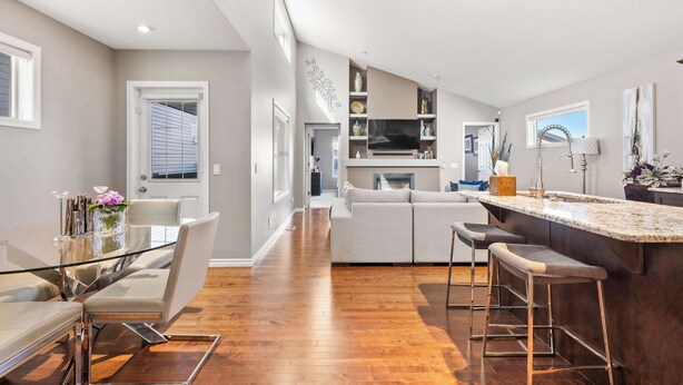 A modern kitchen with a central island, a dining area with a glass table, and a living area with a sofa and bar stools.