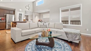 A modern living room with a glass coffee table, a beige sectional sofa, and a kitchen in the background.