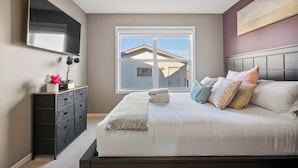 A bedroom with a bed, a dresser, a TV, and a window with a view of a house.